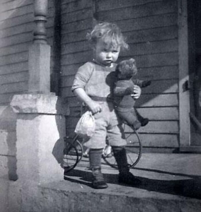 Young child from decades ago holding a teddy bear, standing in front of an old wooden house, illustrating historical change.