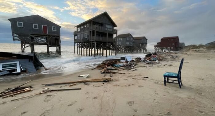 Casas sobre pilotes en la playa con escombros alrededor al atardecer, ejemplo de arquitectura fracasada.