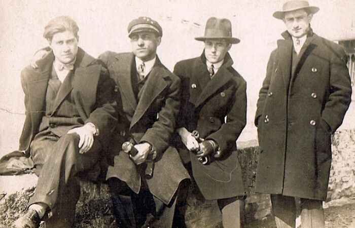 Four men in vintage suits and hats, seated outdoors, showcasing fashion from 50-100 years ago.