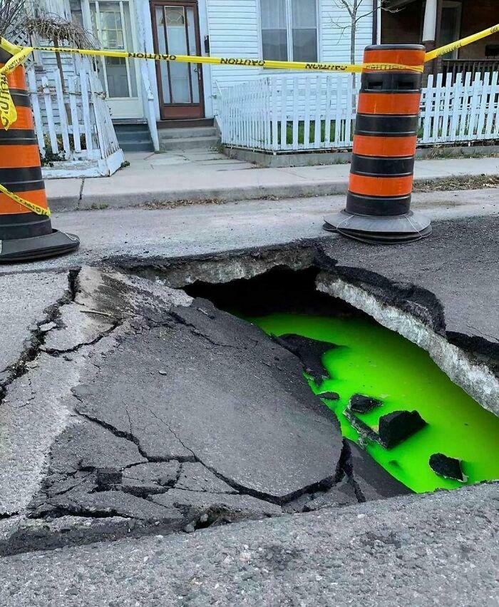 Large road sinkhole filled with bright green liquid marked with caution tape and barrels