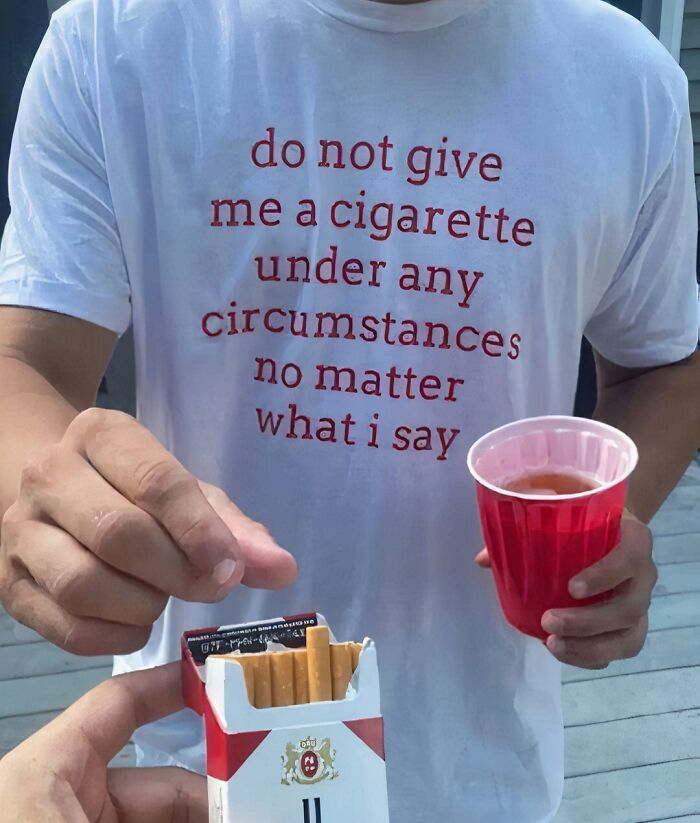 Man wearing a chaotic T-shirt with humorous text, holding a red cup, while being offered cigarettes.