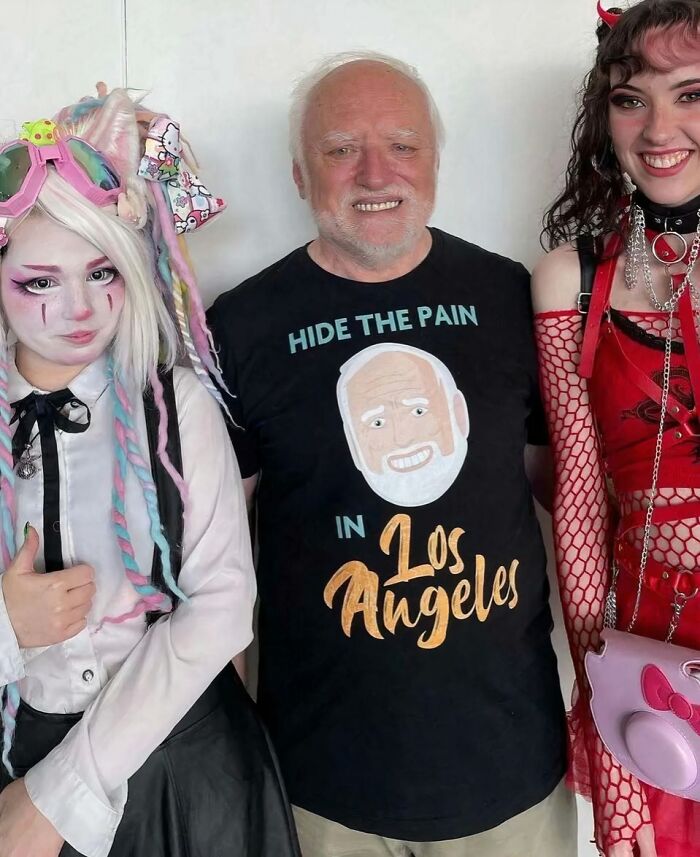 Man wearing chaotic T-shirt with "Hide the Pain in Los Angeles" design, smiling between two colorfully dressed individuals.