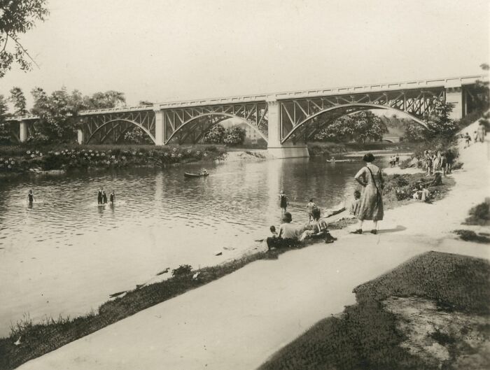 Historic photo of a riverside scene with a bridge, people relaxing by the water, and swimmers, showcasing how much the world has changed.