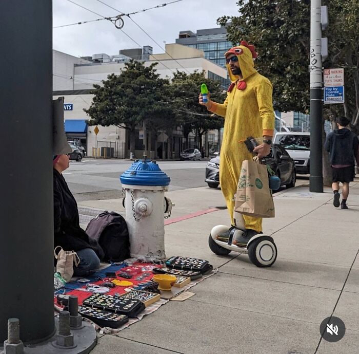 Person in chicken costume on a scooter with a drink, near a street vendor in San Francisco.