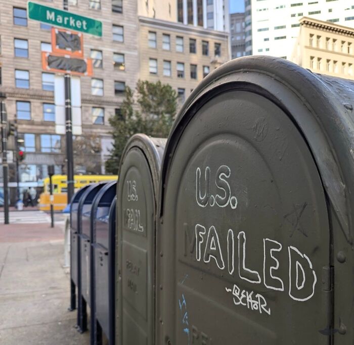 Mailbox with graffiti reading "U.S. Failed" on a San Francisco street corner near Market sign.