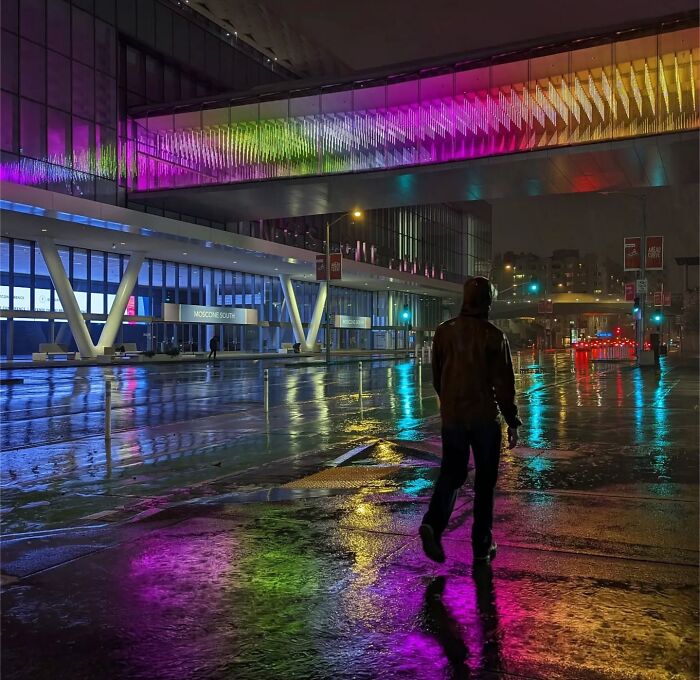 A person walks through a rain-soaked San Francisco street at night, with colorful lights reflecting on the wet pavement.
