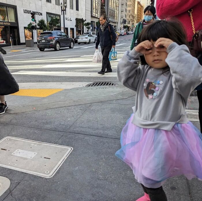 Child in a tutu makes a playful gesture in busy San Francisco street, capturing the city's essence through a candid moment.