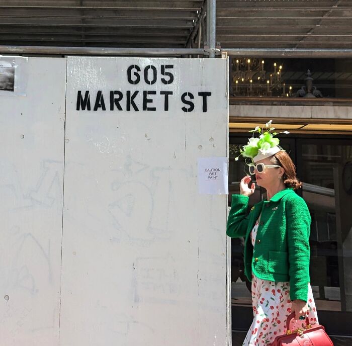 Woman in floral hat and green jacket walking past sign at 605 Market St, San Francisco.
