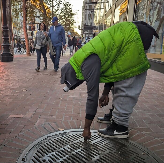 Person in a green vest bending over a manhole cover on a busy San Francisco sidewalk.