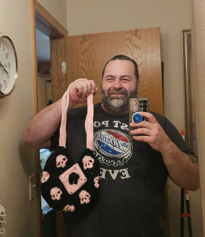 Man smiling and holding a black and pink crocheted bag, showcasing a creative piece made by crochet enthusiasts.