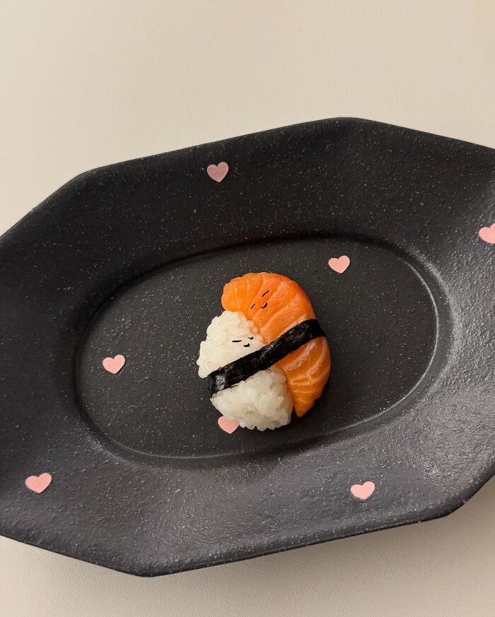 Adorable sushi art by Korean artist, featuring rice and salmon decorated with tiny hearts on a black plate.