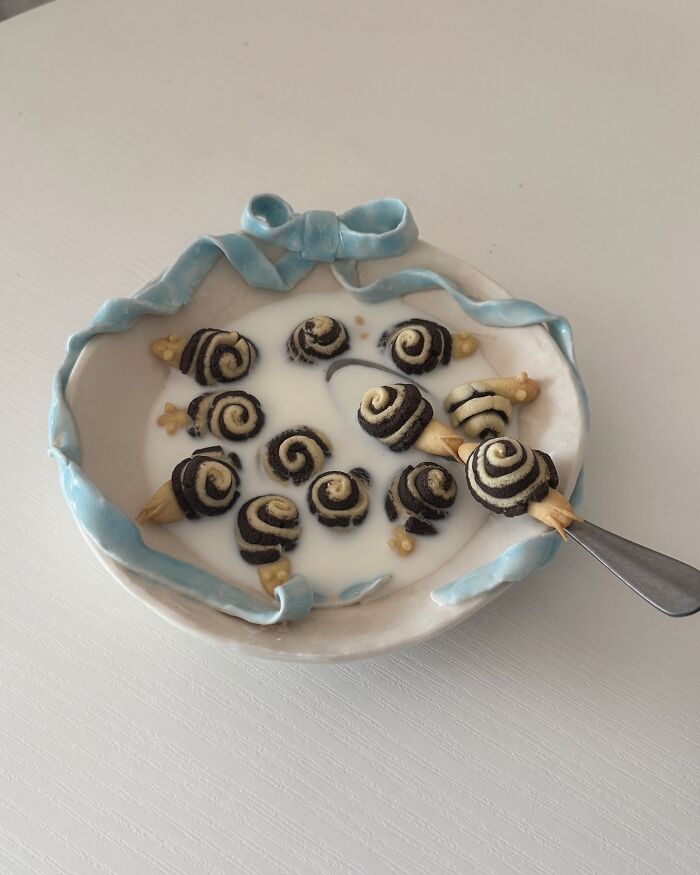 Adorable food art by Korean artist, featuring snail-shaped pastries in milk within a decorative blue-ribbon bowl.