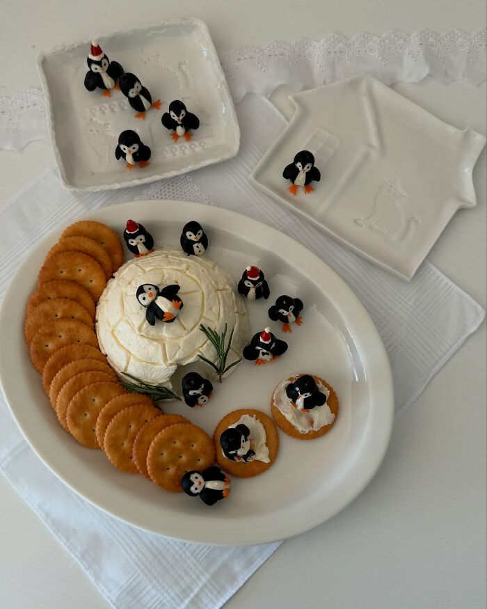 Adorably arranged food with penguin-themed decorations and crackers on a white platter.
