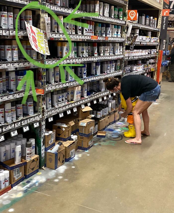 Customer looking at spray paint in a hardware store aisle, with sign labeled "Spray" highlighted in green.