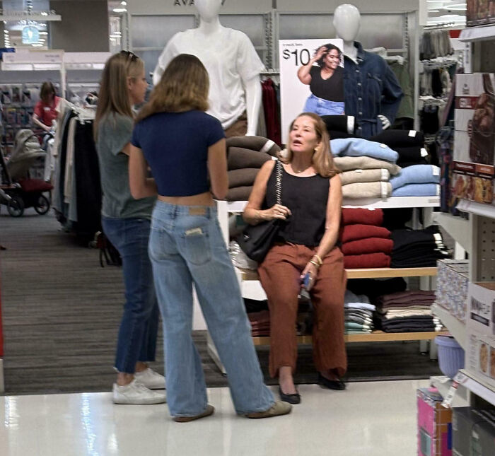 Women talking in a clothing store, displaying customer cluelessness amidst folded sweaters and discount signs.