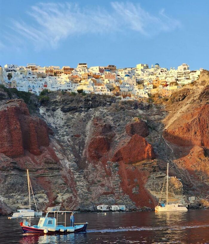 Santorini's iconic cliffside houses illuminated by the sun, with boats sailing on the Aegean Sea.