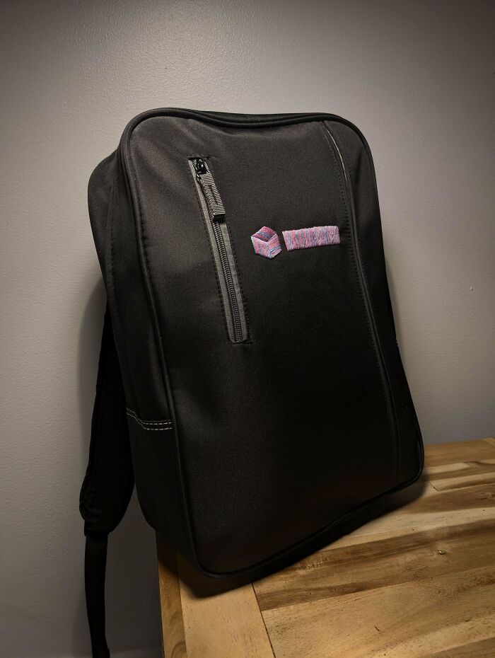 Black backpack with a front zipper pocket, embroidered logo, placed on a wooden surface highlighting consumerism products.