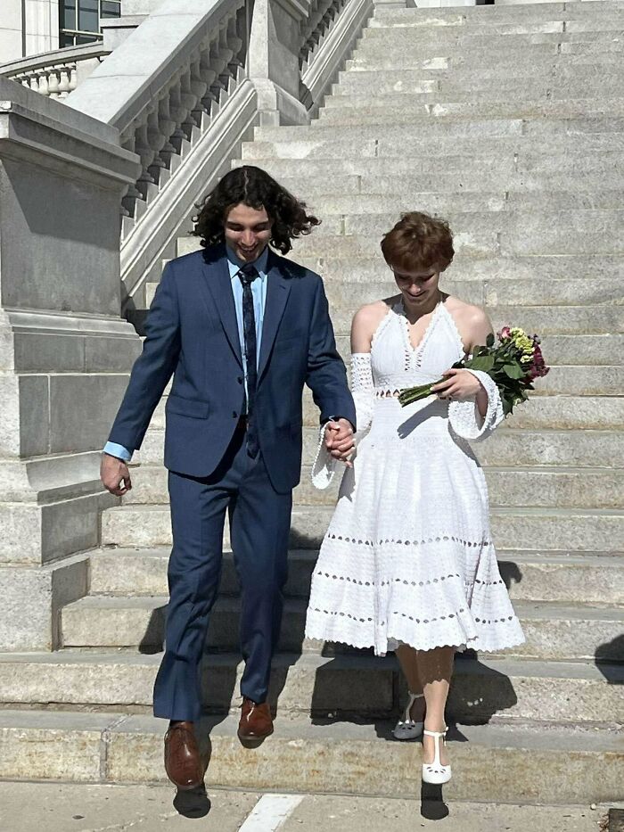 Couple walking down stone steps, woman wearing a white crochet dress holding flowers, man in a blue suit holding her hand.