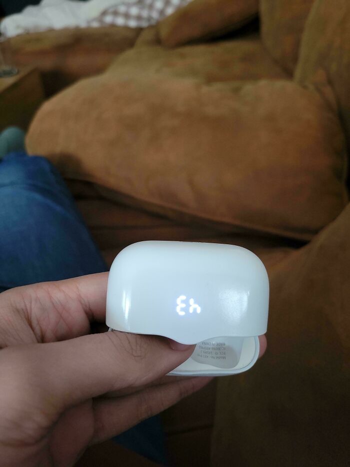A hand holding a plain white charging case in front of a brown couch.
