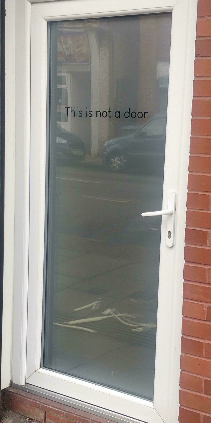 Glass door labeled "This is not a door," reflecting cars and street, humorously highlighting something uninteresting.