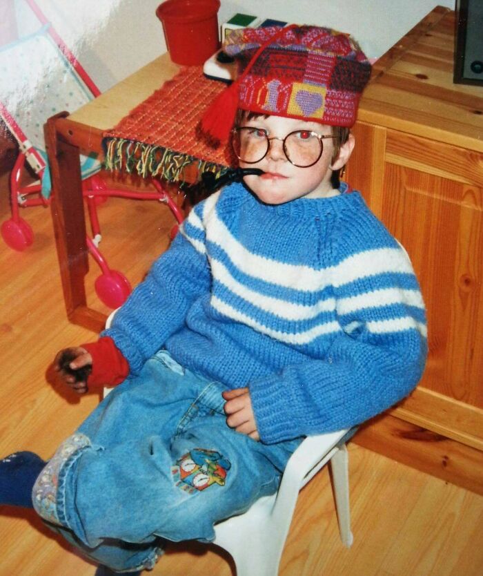 Child in a colorful hat and large glasses wearing a blue striped sweater and patched jeans in a regrettable past photo.