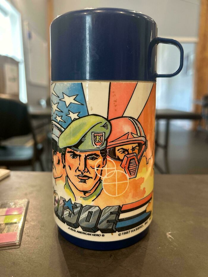 1980s thermos featuring G.I. Joe artwork with soldiers and patriotic motifs on a table.