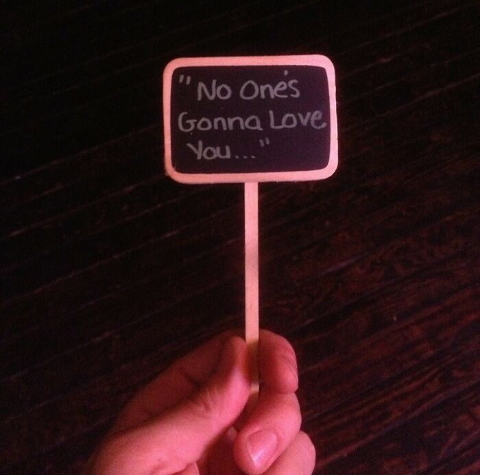 Hand holding a mini sign with a funny passive-aggressive message: "No One's Gonna Love You..." on a dark background.