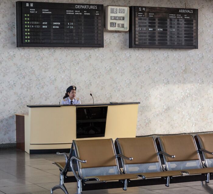 Airport terminal with iconic departure board and staff at desk, vacant seating area visible in foreground.