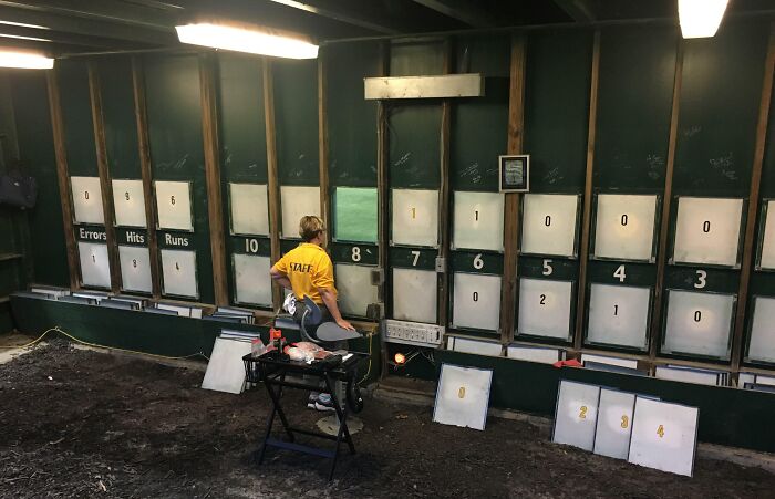 Person in yellow staff shirt managing vintage scoreboard for errors, hits, runs at an iconic place.