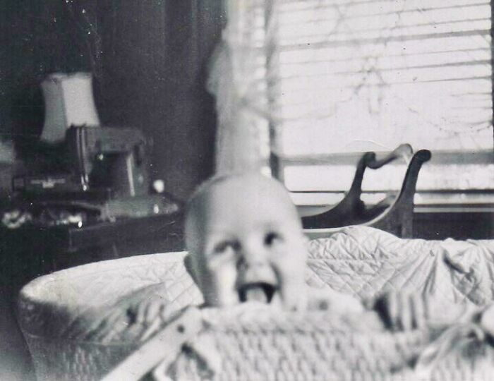 Black and white candid glimpse into the past showing a joyful baby in a vintage bassinet near a sewing machine and window.
