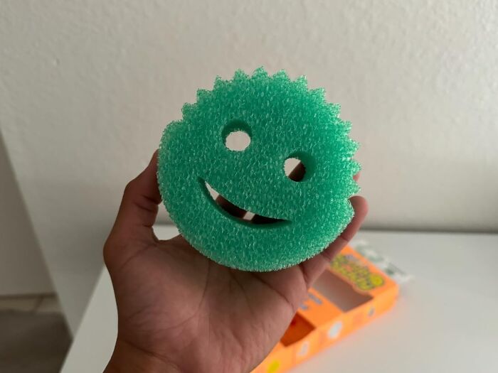 A hand holding a smiley face dish sponge, demonstrating a frugal money-saving tip.