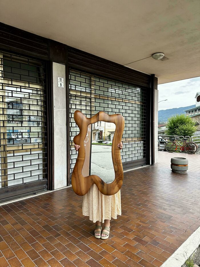 Person holding a unique wooden framed mirror thrift store find standing outside on tiled pavement near closed storefront.