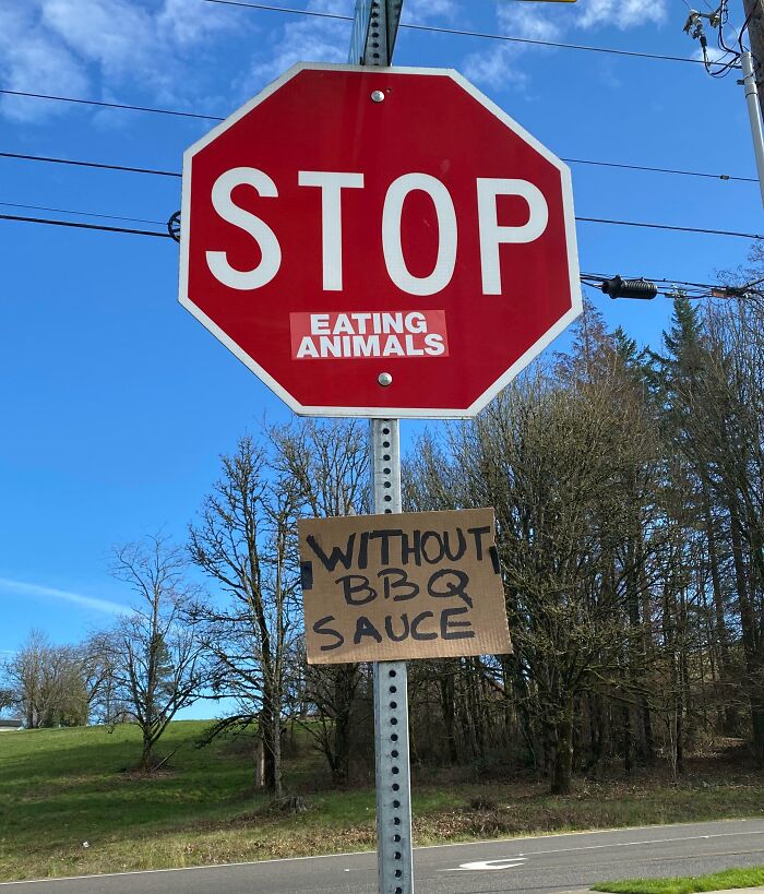 Stop sign with "Eating Animals" sticker and "Without BBQ Sauce" sign. Funny passive-aggressive message in rural setting.