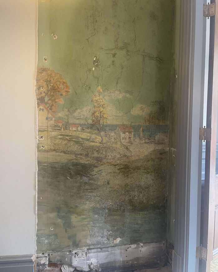 Aged wall mural found in an old house, depicting a serene countryside scene with trees and houses by the water.