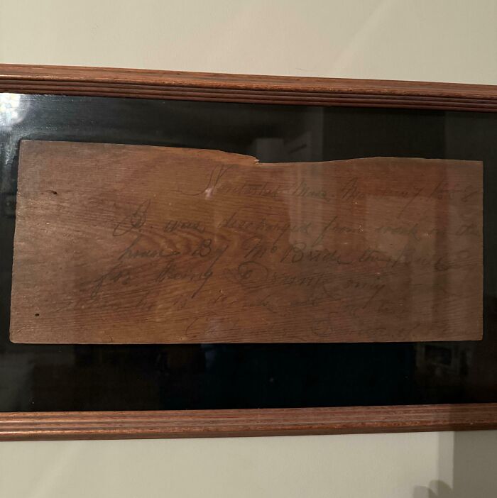 Old wooden artifact with faded handwritten text, displayed in a dark wooden frame.