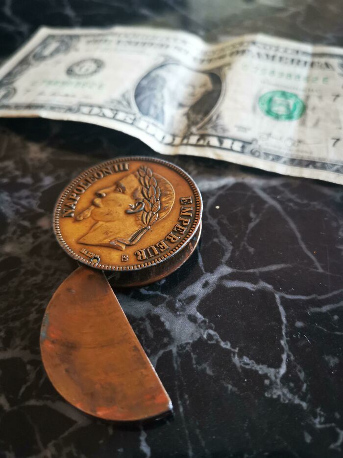 Antique artifact from the past, a copper Napoleon III coin, shown alongside a US dollar bill on a marble surface.