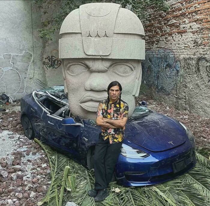 Man standing in front of a crushed blue car and large stone sculpture, highlighting brutal reminders of consumerism impact.