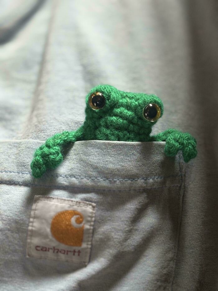 Crochet enthusiast-made small green frog peeking out of a grey Carhartt pocket with large shiny eyes.