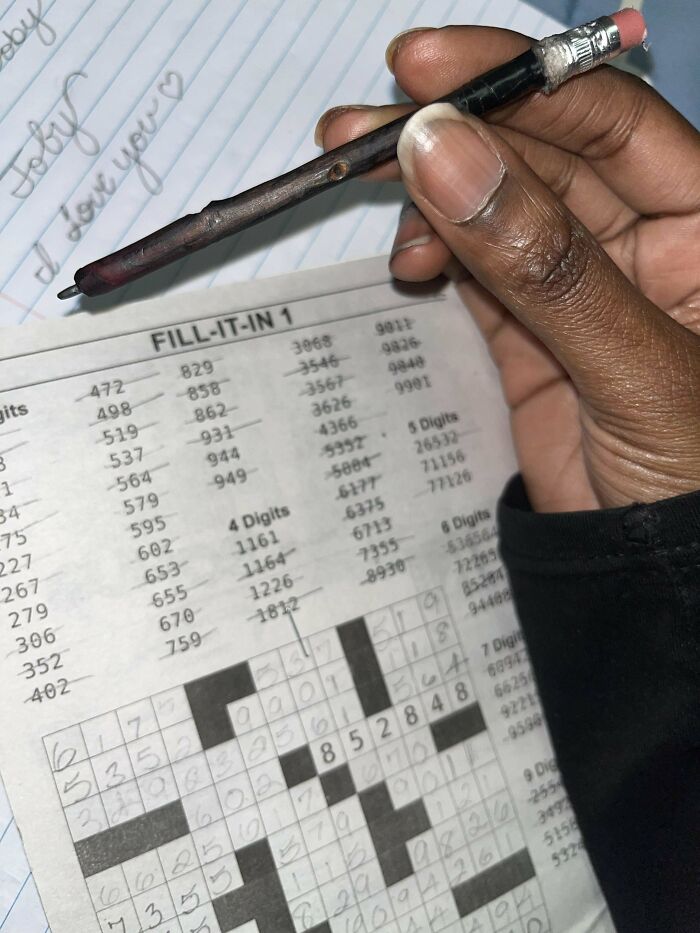 Hand holding a pencil made from a twig, writing on a crossword puzzle page, showcasing a DIY disaster project.