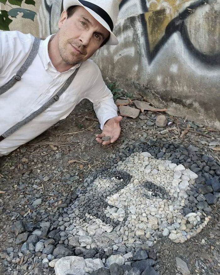 British artist posing with intricate pebble mosaic portrait beside a graffiti wall.