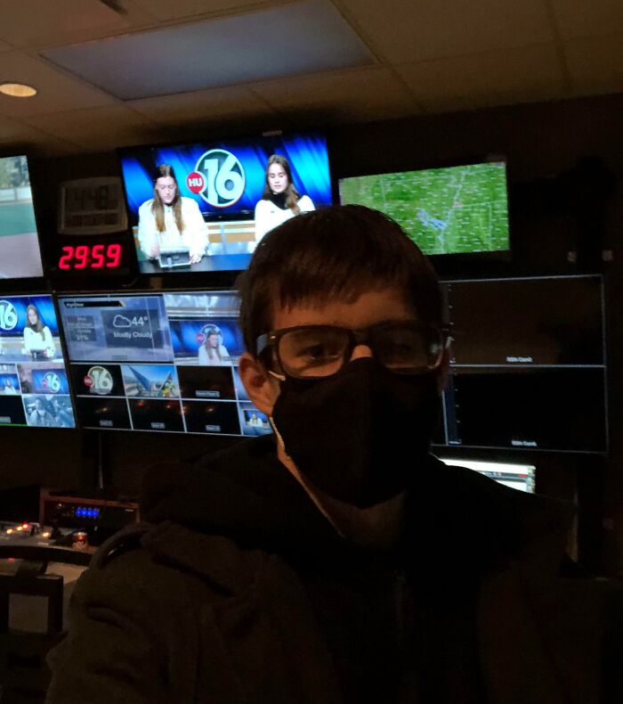 Person in a studio control room, acting like they belong, with monitors displaying broadcasts and news anchors.