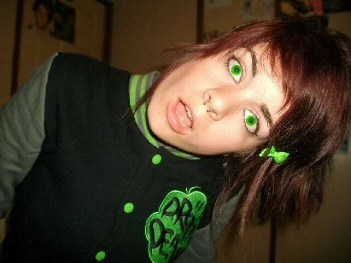 Young person with green contact lenses and nose piercing, wearing a black and green jacket, reflecting a regrettable past photo.
