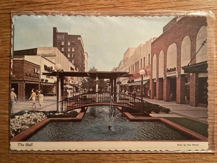 Vintage mall scene with a water fountain and bridge, surrounded by storefronts and people strolling.