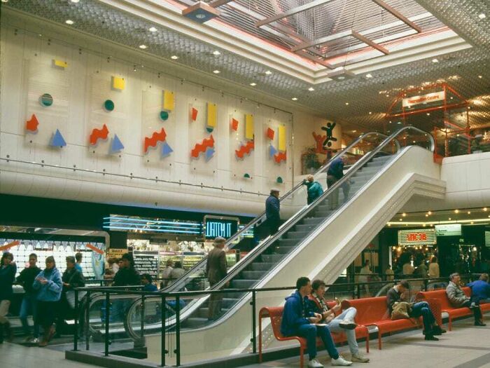 Vintage mall scene with an escalator, colorful wall art, and people in a bustling food court area.