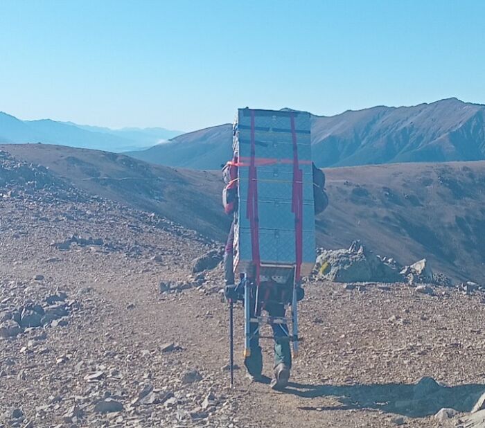 Hiker balancing a large, strapped box on rugged mountain trail, embodying "epic airline baggage fail" concept.