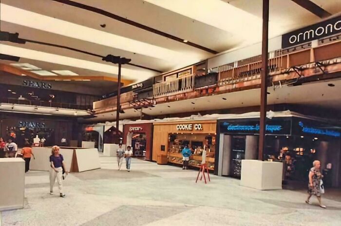 Vintage mall scene with people walking near Sears and Cookie Co., evoking a nostalgic food court atmosphere.