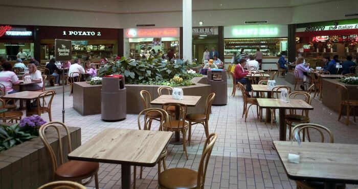 Vintage mall food court with wooden tables, chairs, and people dining, surrounded by food outlets and plants.