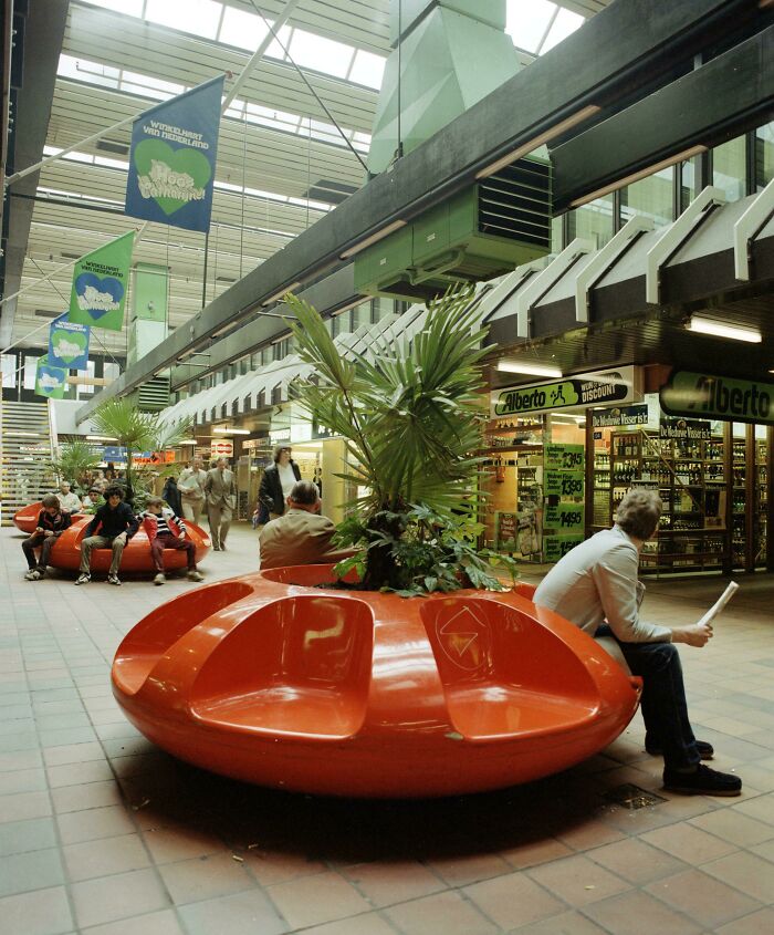 Vintage mall scene with shoppers and retro seating near a food court area.