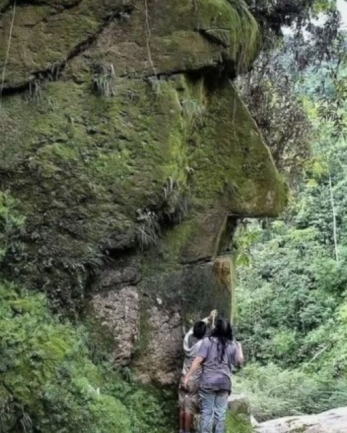 Two people examining an ancient rock formation resembling a face, covered in moss and greenery.