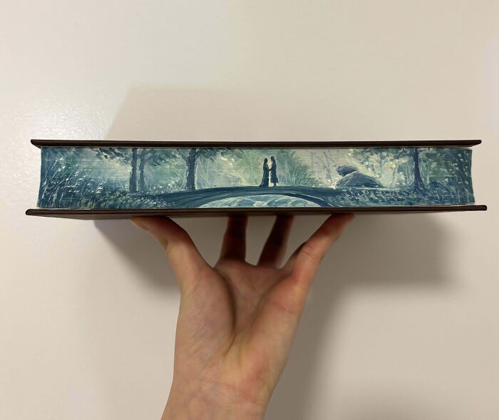 Hand holding a book with a detailed scenic painting on the edges, showcasing a DIY project that looks amazing.